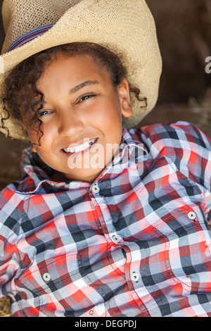 Beautiful happy mixed race African American female girl child wearing straw cowboy hat & plaid shirt sitting in hay filled barn Stock Photo