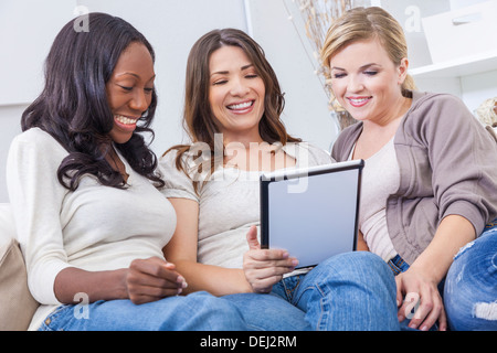 Three beautiful young women at home sitting on sofa or settee using a tablet PC computer and smiling Stock Photo