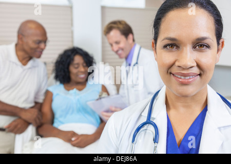 Happy woman female Hispanic Latina Doctor in hospital with patient recovering in hospital bed & male doctor in background Stock Photo