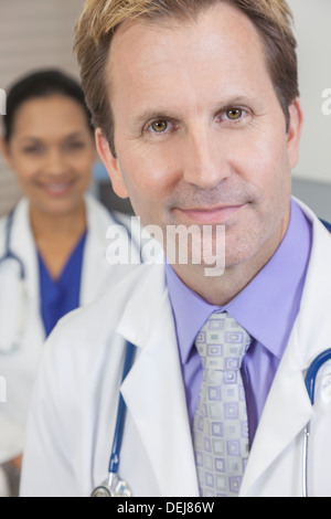 A male doctor wearing a shirt, tie and stethoscope with female Latina colleague behind him Stock Photo