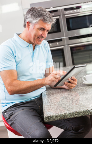 Middle aged man using tablet computer at home in the kitchen drinking coffee or tea Stock Photo