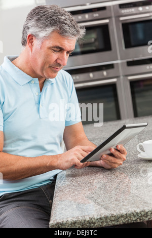 Middle aged man using tablet computer at home in the kitchen drinking coffee or tea Stock Photo
