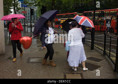 Women, incl one pregnant with a heart-shaped balloon, on a street corner shelter under umbrellas during damp, gloomy weather in central London. Stock Photo