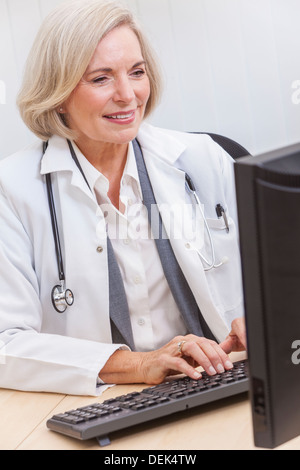 A senior female doctor sitting at a desk in an office with a computer, wearing a suit and stethoscope Stock Photo
