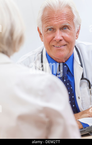 Senior male doctor sitting at a desk in an office with a computer, wearing a shirt, tie and stethoscope seeing a female patient Stock Photo