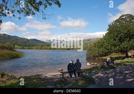Elderly couple people sitting sat on wooden bench seat looking across Elterwater to Langdale Pikes Lake District summer Cumbria England UK Britain Stock Photo