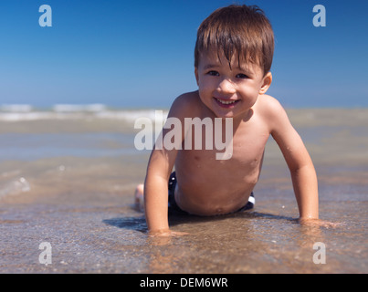 Portrait of a happy smiling three year old boy lying on a sandy shore in the water