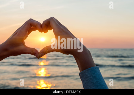 Hands and fingers in heart shape framing setting sun at sunset over ocean Stock Photo