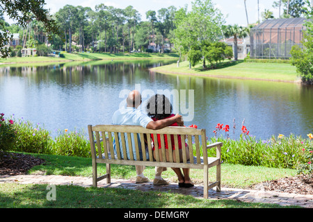 Rear view of a happy romantic senior African American couple sitting on a park bench embracing looking at a lake Stock Photo