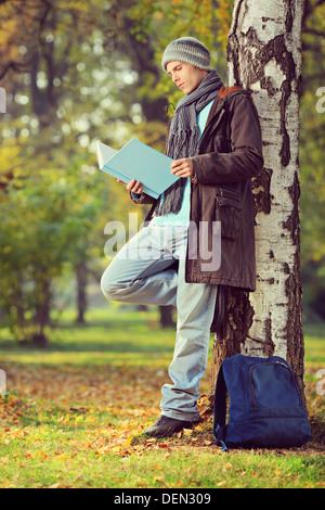 Full length portrait of a male student leaning on a tree and reading a book in a park Stock Photo