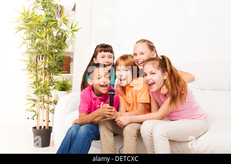 Group of five happy diversity kids, boys and girls, singing together sitting on the coach in living room Stock Photo