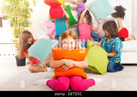 Happy smiling little girl hugging pillow with large group of her friends pillow fighting on the background Stock Photo