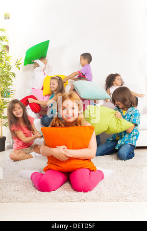 Happy smiling little girl hugging pillow with large group of her friends fighting with pillow on the coach Stock Photo