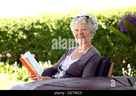 Happy senior woman with book in hand sitting in her backyard looking at camera smiling - Elder woman reading novel in garden Stock Photo