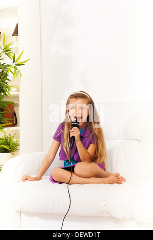 Close portrait of little Caucasian with strong face expression, five years old girl with blond long hair singing and scramming to microphone performing sitting on the coach Stock Photo