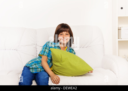 One happy calm and relaxed little boy 6 years old sitting with green pillow on the white leather coach in living room at home Stock Photo