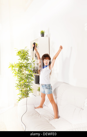 Singing cute little happy girl with microphone and lifted hands both standing on the coach in home living room Stock Photo