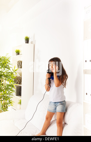 Singing cute little happy girl singing to microphone s both standing on the coach in home living room Stock Photo