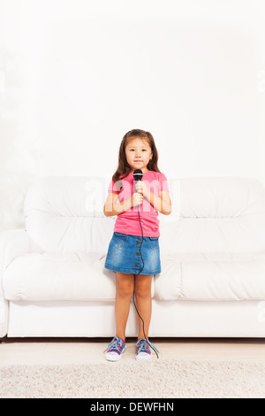 Cute little Asian five years old girl with dark long hair singing to microphone performing standing in living room Stock Photo