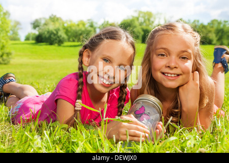 Close-up of two happy little nine years old girls hold jar with butterfly laying in the grass and showing big smile on happy faces Stock Photo