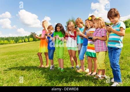 Large group of busy kids, boys and girls looking at their phones texting sms and playing staying outside in the field on bright sunny summer day Stock Photo