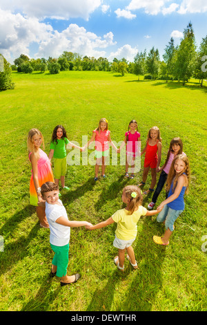 Large group of happy girls play roundelay and stand in circle in the park on the green grass on sunny summer day, view from above Stock Photo