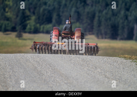 Agricultural scene of farmer towing, machinery, a discer, with a old tractor, on a rural country road back to homestead farm. Stock Photo