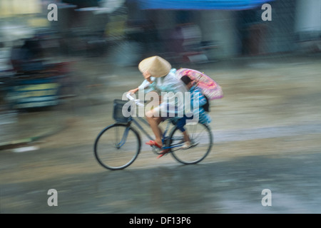 Woman in a conical hat riding a bicycle through a rainstorm, with her daughter holding an umbrella behind, Hoi An, Vietnam Stock Photo