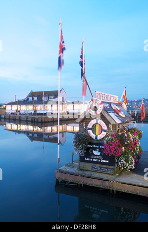 Flowers and flags decorate the ticket hut at West Bay River Boat Hire. Across the water is the Riverside Restaurant. Dorset, England, United Kingdom. Stock Photo