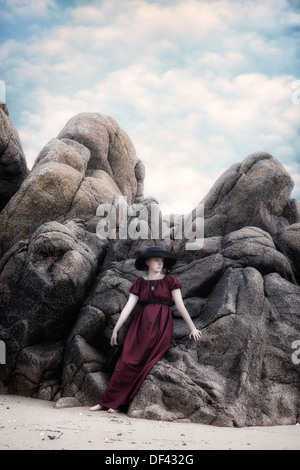 a young girl in a red dress with a black sunhat is leaning against rocks at the beach