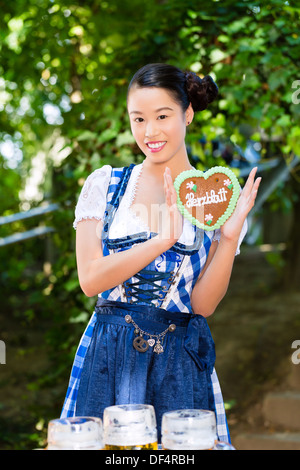 Young Asian woman in traditional Bavarian clothes or tracht with a gingerbread souvenir heart in beergarden on Oktoberfest Stock Photo