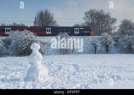 Train travelling on cold winter day, along railway line, passing single snowman standing in snow-covered field - Burley in Wharfedale, England, UK. Stock Photo
