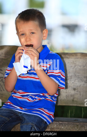 Happy childhood. Boy child or kid sitting on bench in summer park eating sweets chocolate Stock Photo