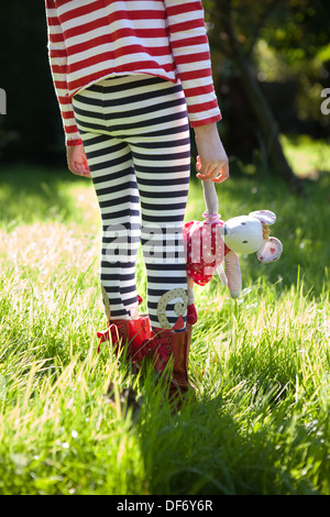 Child wearing stripes, standing in the long, overgrown, green grass of a meadow and holding a toy mouse for comfort. Stock Photo