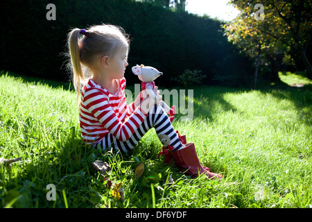 Little blond girl sitting in the long, overgrown green grass of a meadow on a dry summer day and holding a toy mouse for comfort Stock Photo