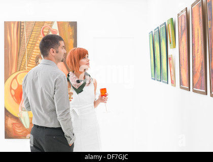 young caucasian couple standing in a gallery and holding colorful glasses of wine in their hands, contemplating artwork displayed on walls Stock Photo