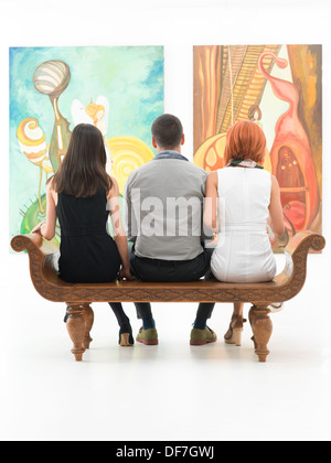 back view of three young people sitting on a wooden bench in a museum admiring large paintings Stock Photo