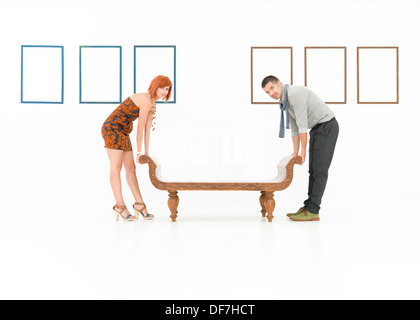 two caucasian people trying to lift up a wooden bench in a white room with empty frames displayed on walls Stock Photo