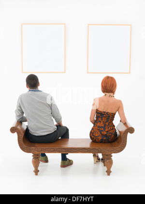rear view of man an woman sitting on a wooden bench looking at empty frames displayed on a white wall in front of them Stock Photo