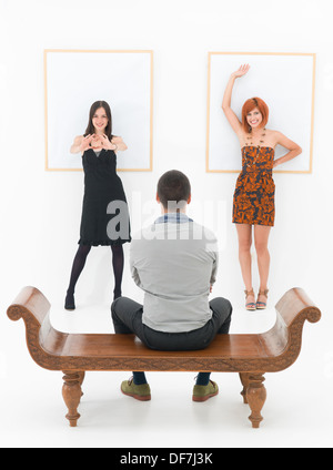 two happy women leaning angaist a white wall with empty frames in front of a man sitting on a bench, looking at them Stock Photo