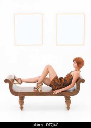 side view of beautiful woman resting on a wooden bench in front of two large empty frames on a white wall Stock Photo