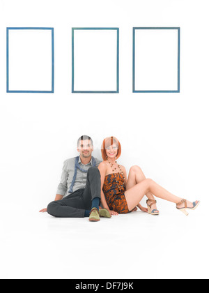 front view of happy man and woma resting on a floor against a whit wall withe empty blue frames Stock Photo