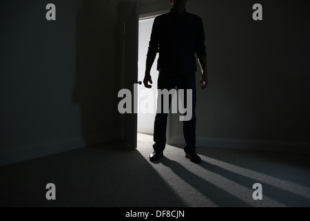 Shadowy figure of a man standing by an doorway leading to a dark room ...