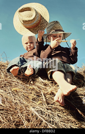 Two young children, a little boy and his baby brother are sitting on a hay bale, wearing straw hats, in front of a blue sky Stock Photo