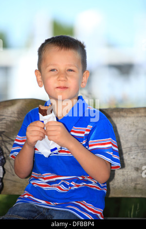 Happy childhood. Boy child or kid sitting on bench in summer park eating sweets chocolate Stock Photo