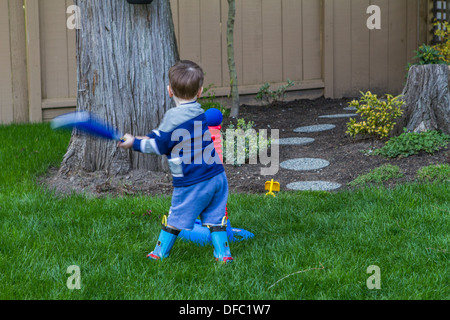 Model released three year old boy, swinging blue plastic bat, as he plays t-ball on the green grass of his back yard. Stock Photo