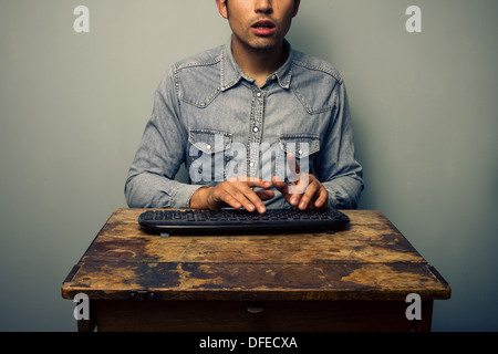 Young man is sitting at an old wooden desk and typing on a wireless keyboard Stock Photo