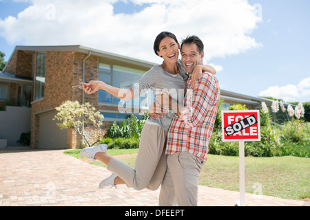 Portrait of enthusiastic couple hugging outside house with For Sale sign Stock Photo