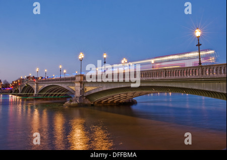 A bus travels over Battersea bridge whilst a boat travels underneath on the River Thames at dusk Stock Photo