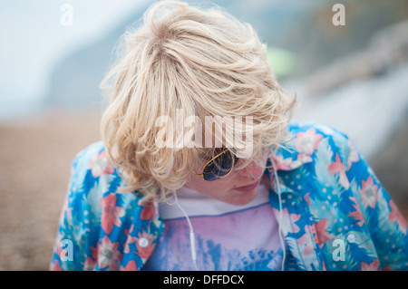 Cool long blond-haired teenage boy wearing Raybans listening to music on his mp3 player sitting on a beach Stock Photo